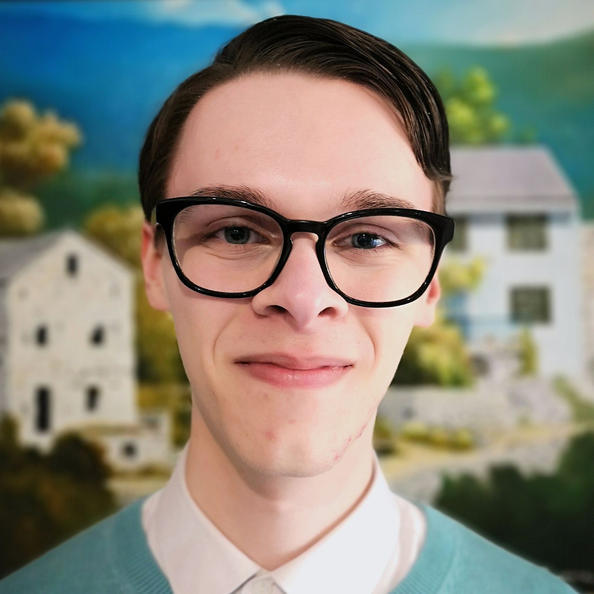 Image of Julian with brown hair, black glasses,
a white shirt, and blue jumper standing in front
of a scenery painting
